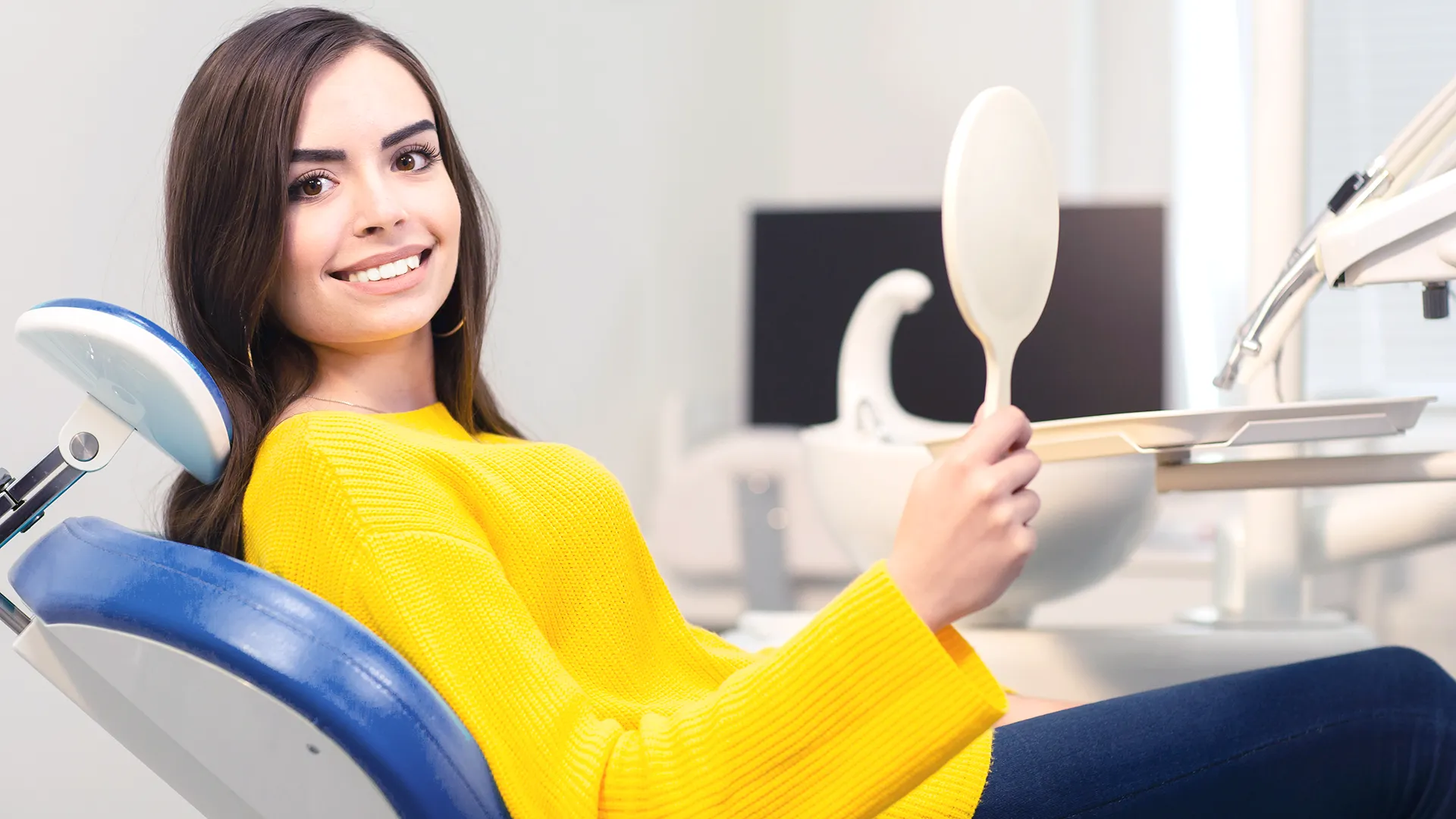 Mujer joven con un suéter amarillo brillante sentada en un sillón dental, sonriendo mientras examina sus dientes en un espejo de mano, en un consultorio dental moderno.