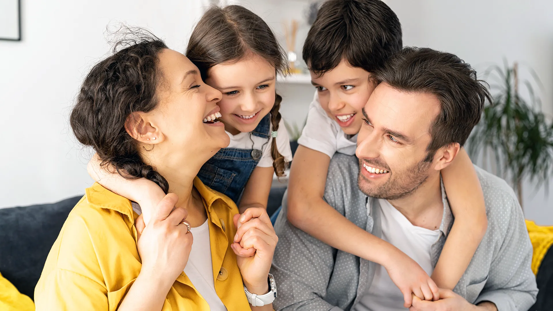 Padres sentados sonriéndose mutuamente, con su hija en la espalda de la madre y su hijo en la espalda del padre.
