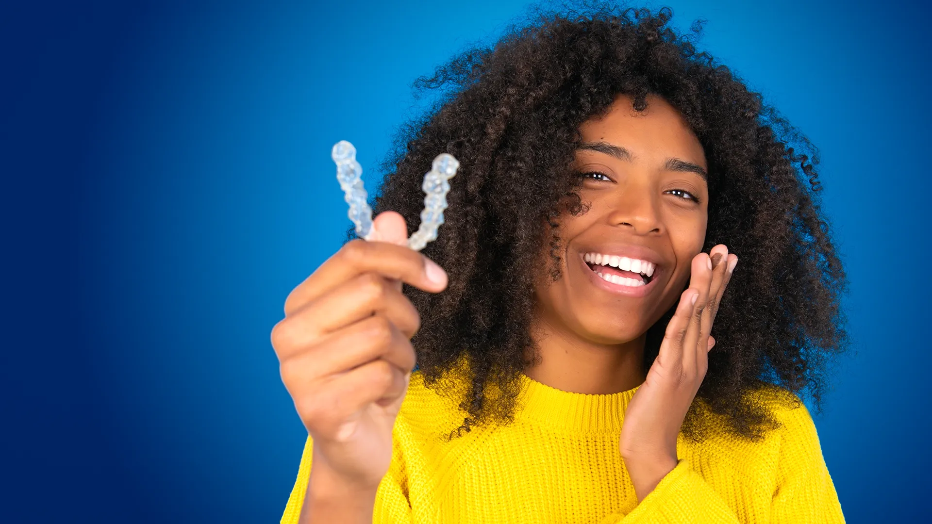 Mujer sonriendo frente a un fondo azul mientras sostiene alineadores transparentes.