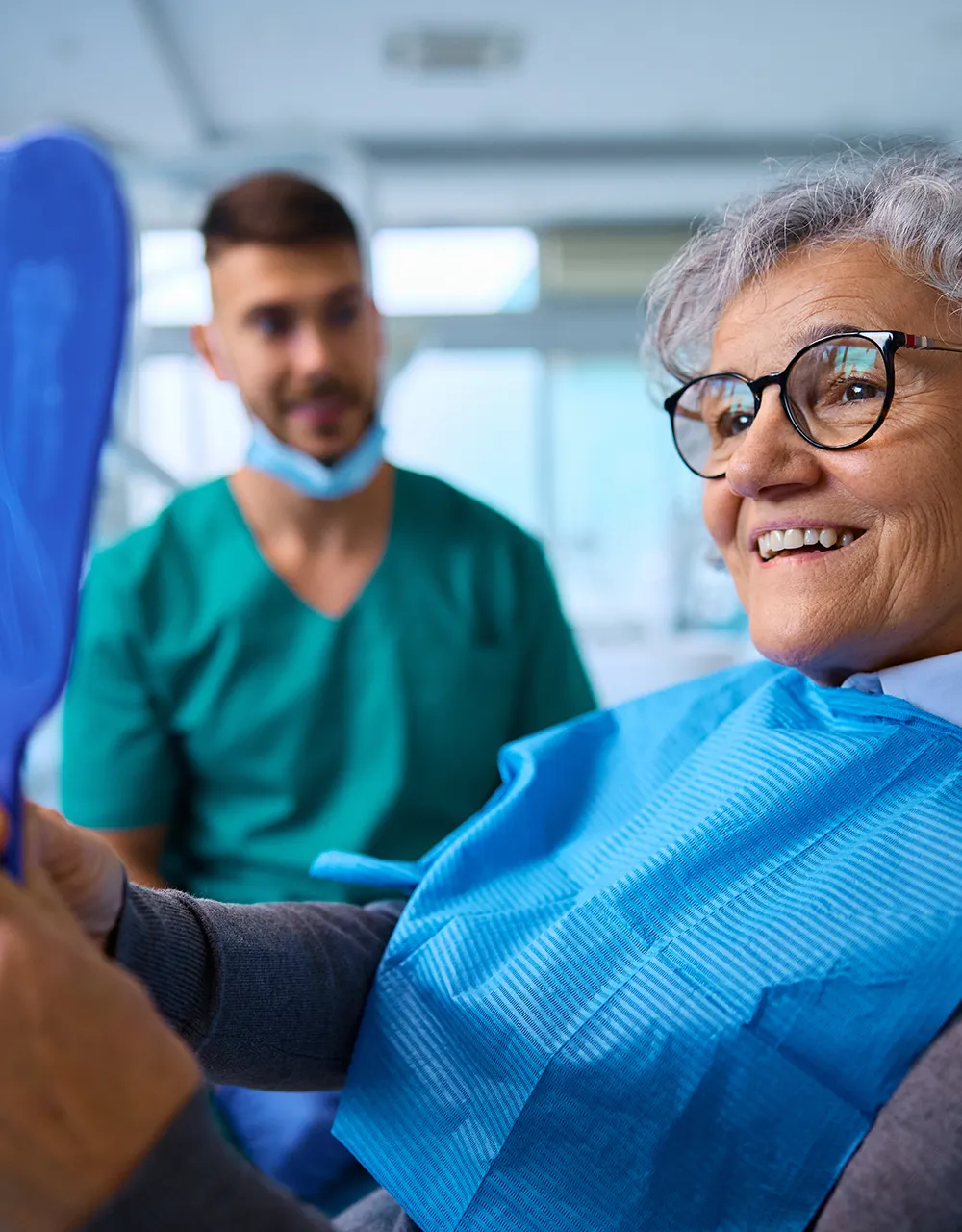 Mujer mayor sonriente con gafas sosteniendo un espejo, examinando sus nuevas dentaduras, con un profesional dental de pie en el fondo.