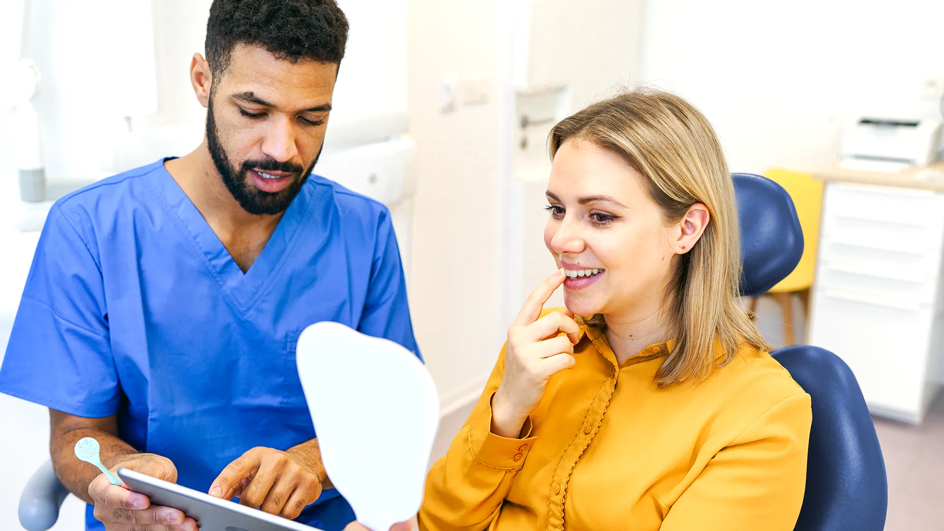 Mujer sentada en el sillón dental señalando su diente en el espejo mientras el dentista le muestra información en una tableta.