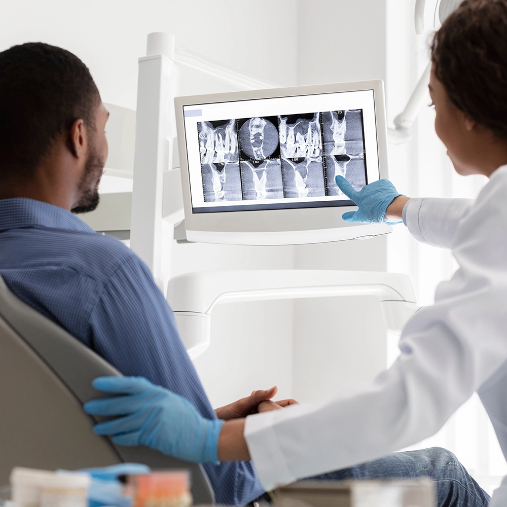 Hombre sentado en el sillón dental mientras el dentista a su lado señala radiografías dentales en una pantalla.