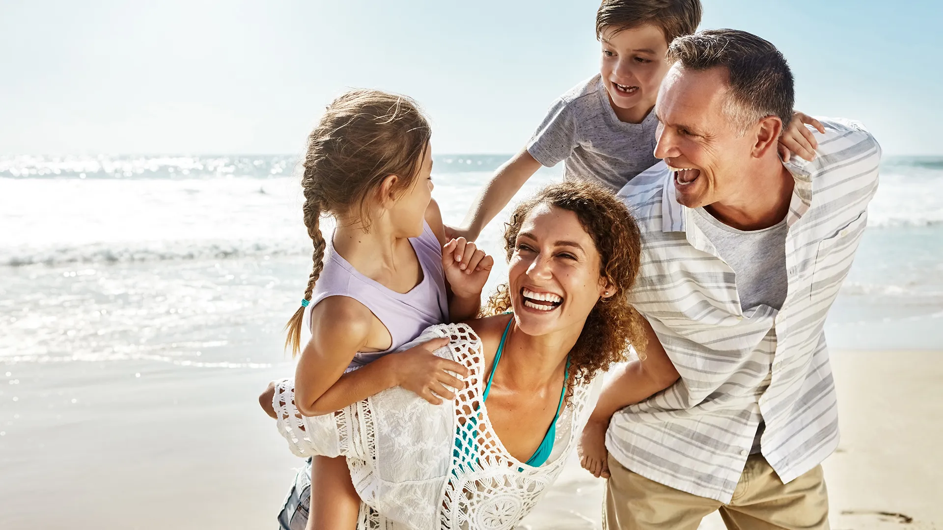 Familia en la playa con los padres llevando a dos niños—una niña y un niño—en sus espaldas, todos riendo.