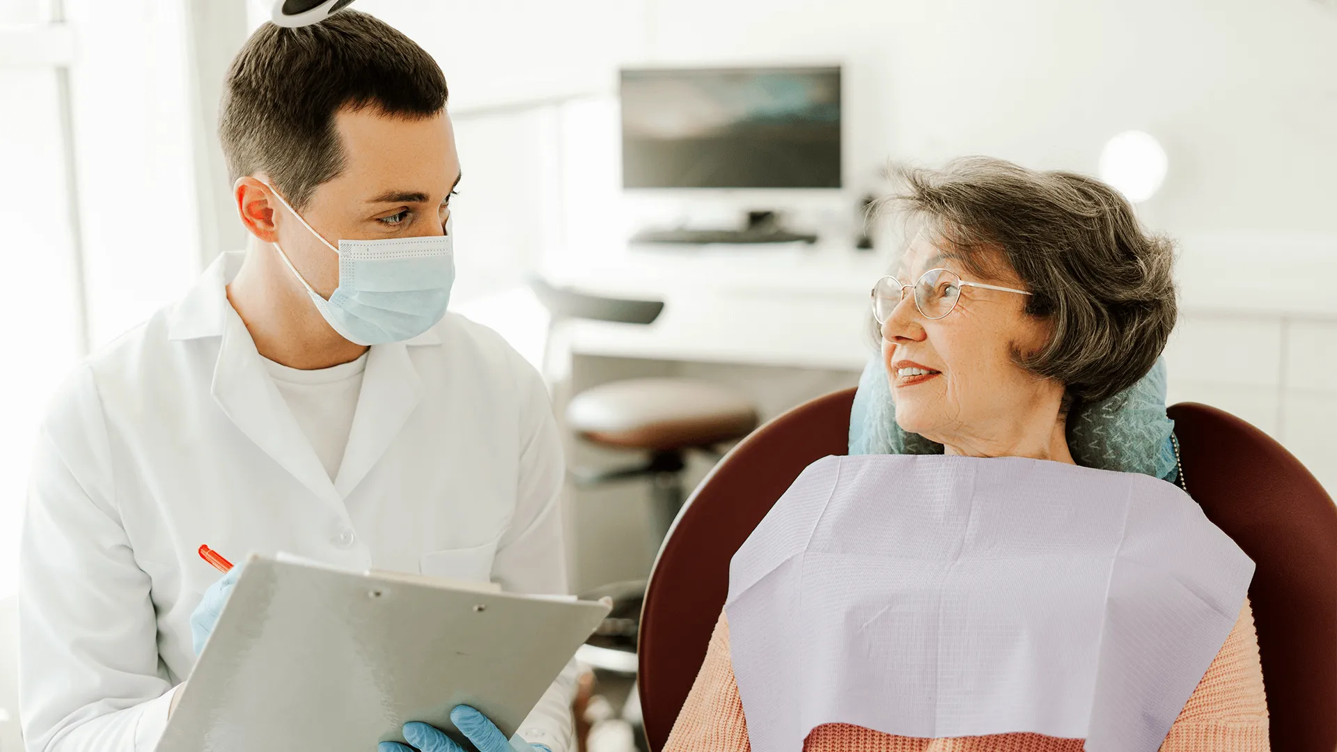 Mujer mayor sentada en el sillón dental mientras el dentista con mascarilla toma notas.