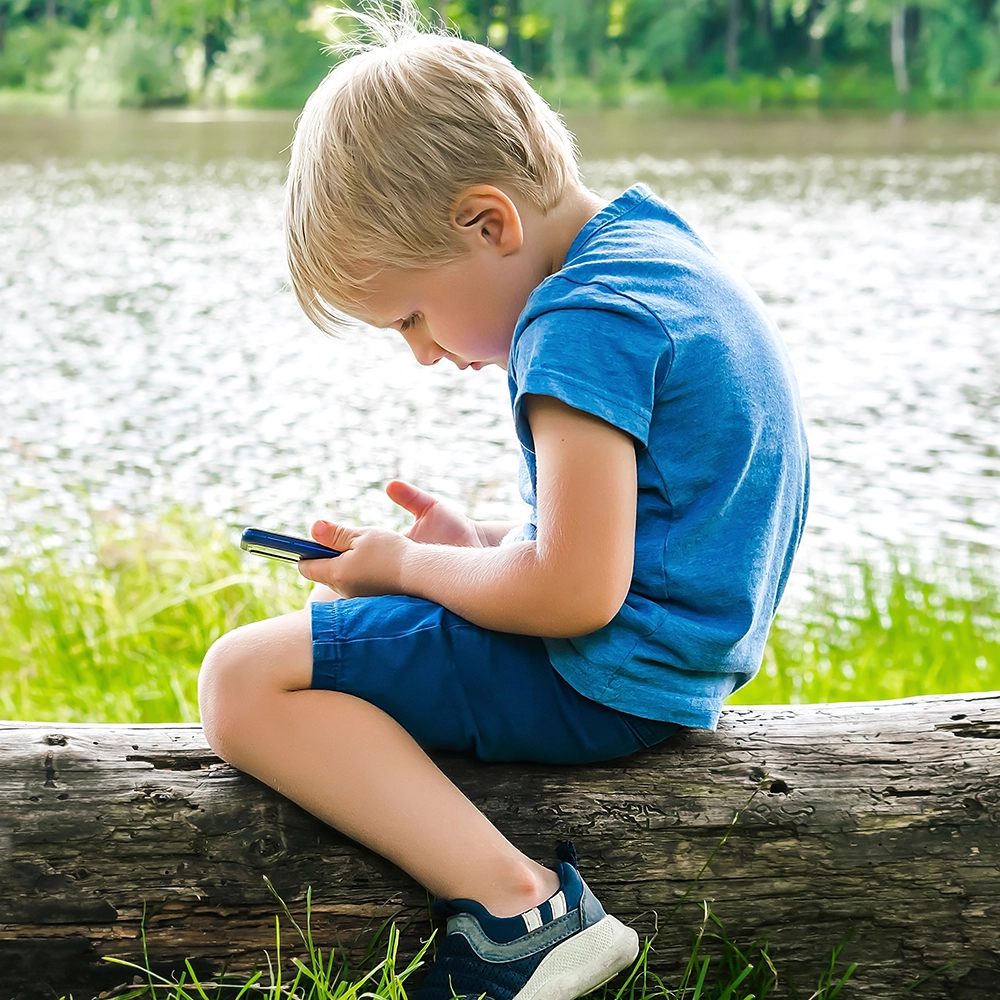 Niño sentado sobre un tronco de madera mirando algo en un teléfono inteligente