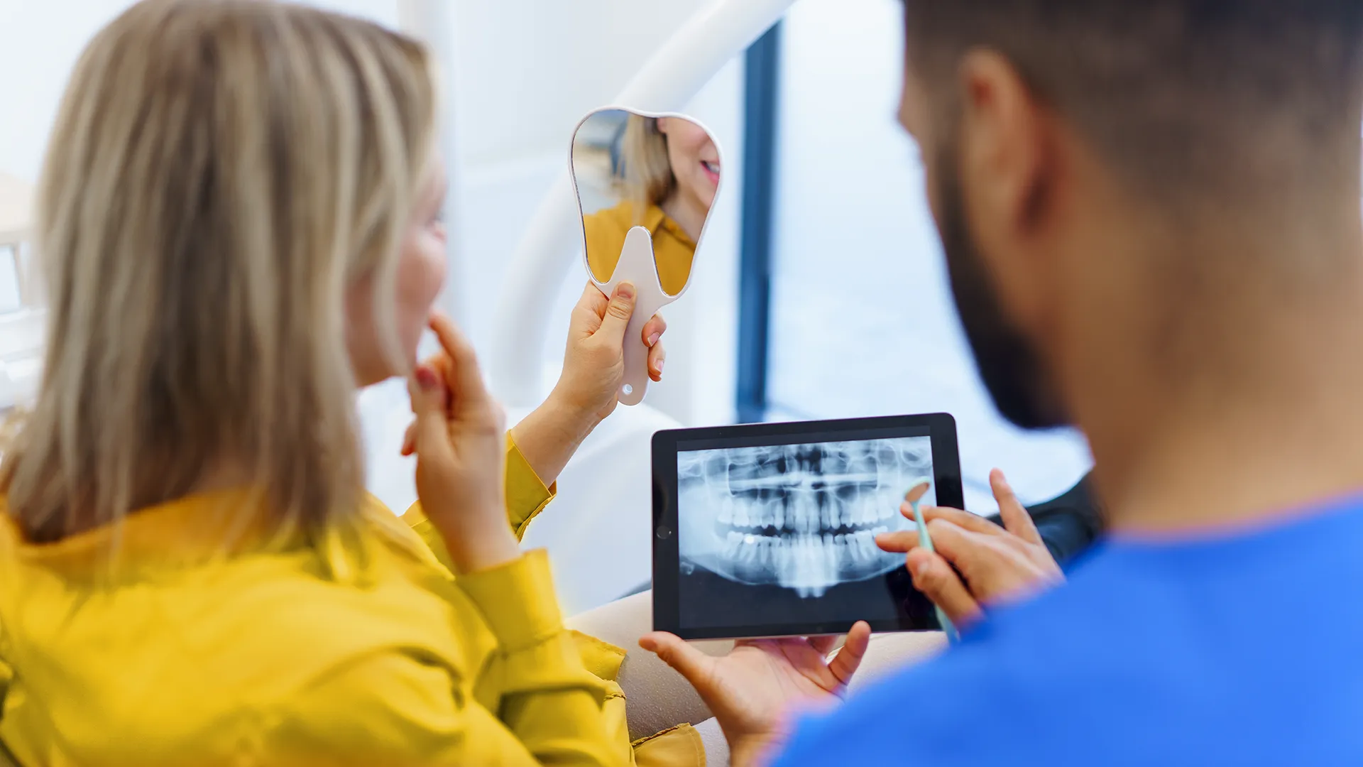 Mujer sosteniendo un espejo para revisar sus dientes mientras el dentista a su lado revisa radiografías dentales en una tableta.