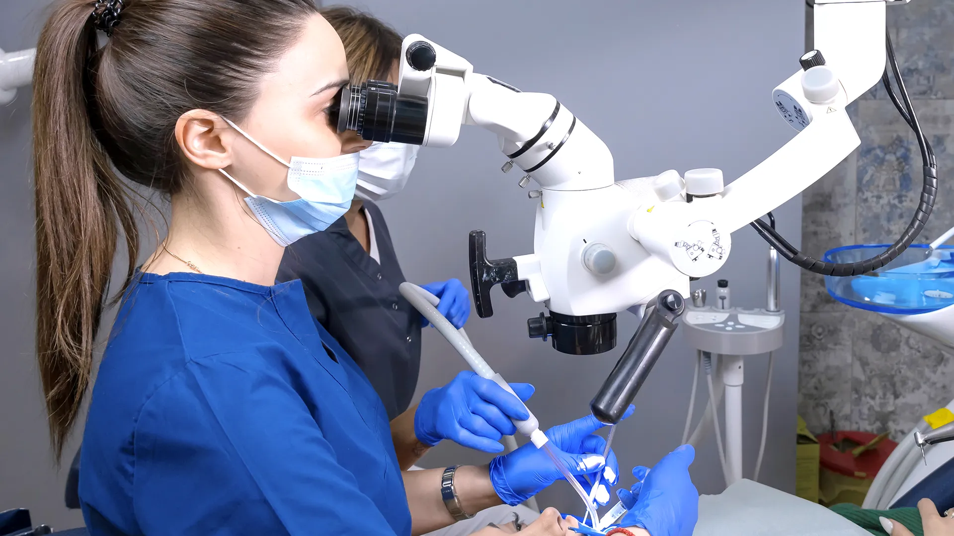 Dentista realizando un procedimiento con herramientas dentales y un microscopio mientras el paciente está recostado en el sillón dental.