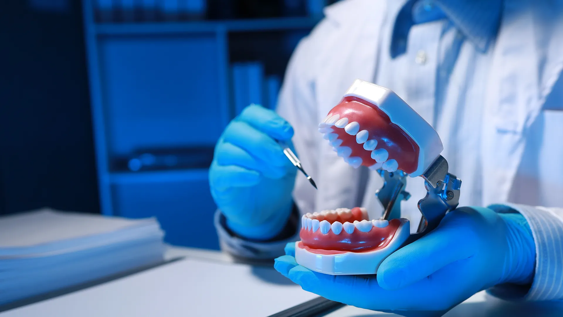 Profesional dental con guantes azules ajustando un juego de dentaduras completas en un modelo, iluminado bajo luz azul para enfatizar la precisión y el detalle en el ajuste de la dentadura.