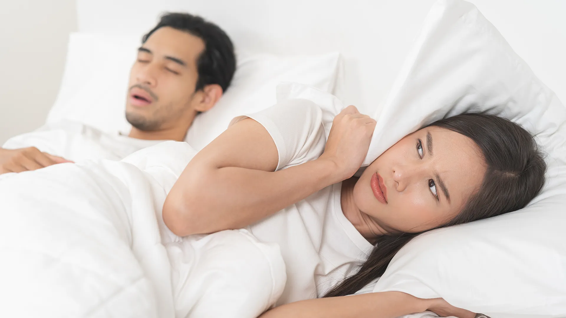 Mujer acostada en la cama con su esposo, cubriéndose los oídos con una almohada debido a sus ronquidos.