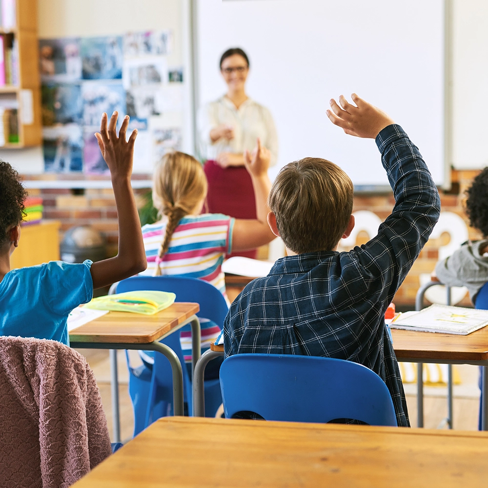Niños sentados en un aula levantando la mano mientras miran al maestro.