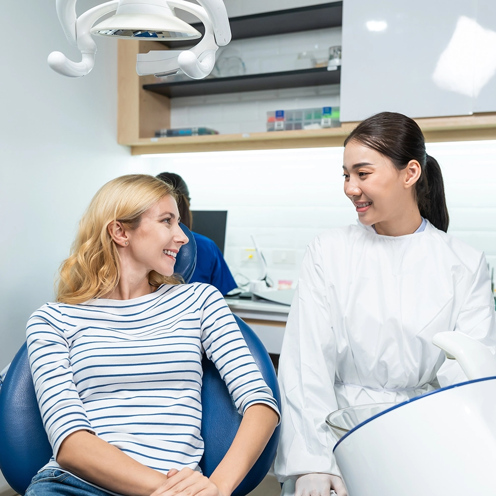 Dentista en bata blanca discutiendo el tratamiento con una paciente rubia sonriente en un sillón dental, mostrando una visita dental cómoda e interactiva.